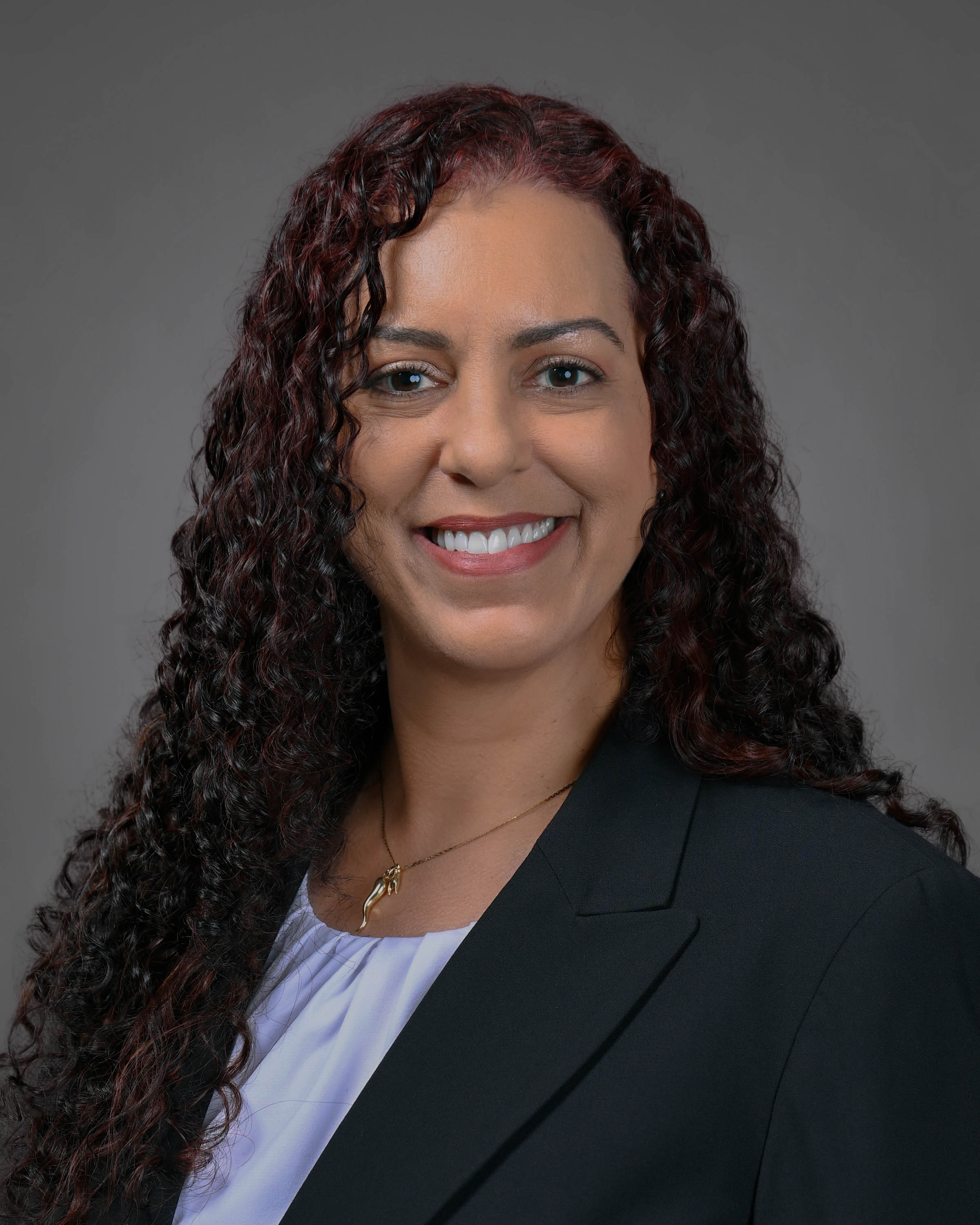 Kind woman smiling with dark brown curly hair wearing a dark jacket with a white shirt.